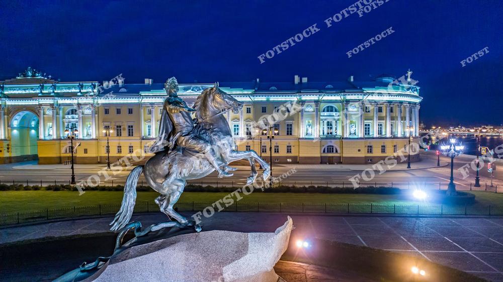 На переднем плане — знаменитый памятник Петру I, известный как Медный всадник, установленный на Сенатской площади. Позади него возвышается здание Сената и Синода, выполненное в стиле классицизма. Площадь красиво подсвечена, а фонари мягко освещают территорию у Невы. Ночная атмосфера подчеркивает торжественность и величие монумента.